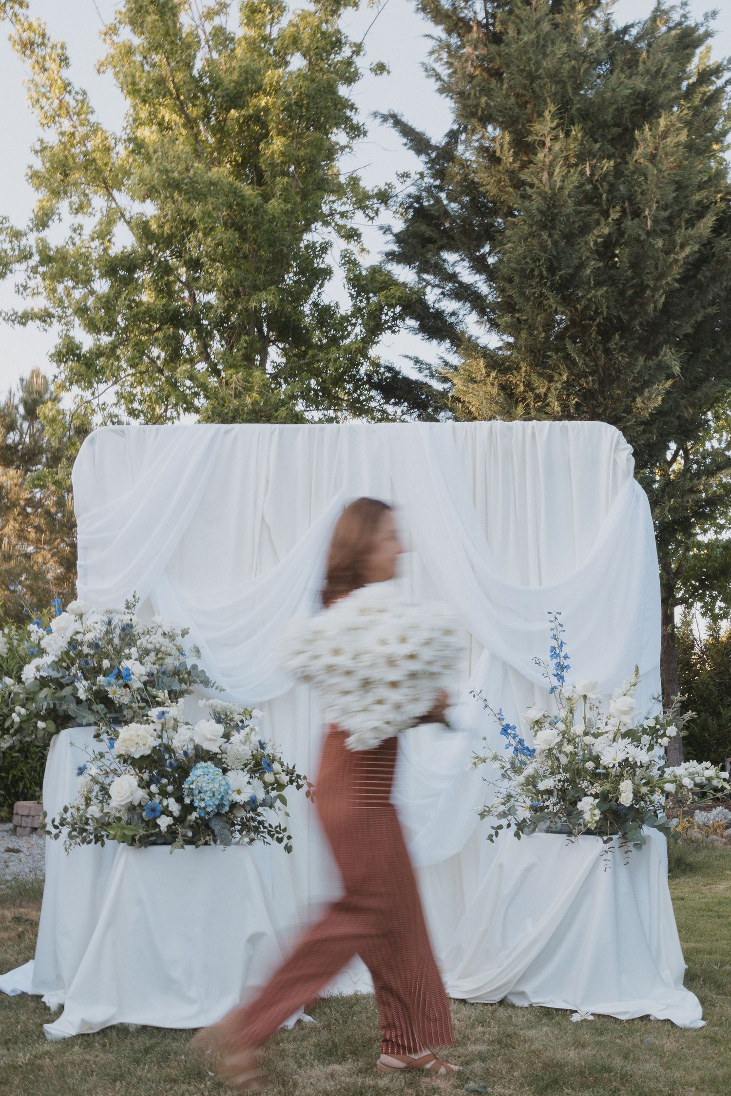White fabric backdrop decorated with blue flowers and greenery for outdoor wedding ceremony.