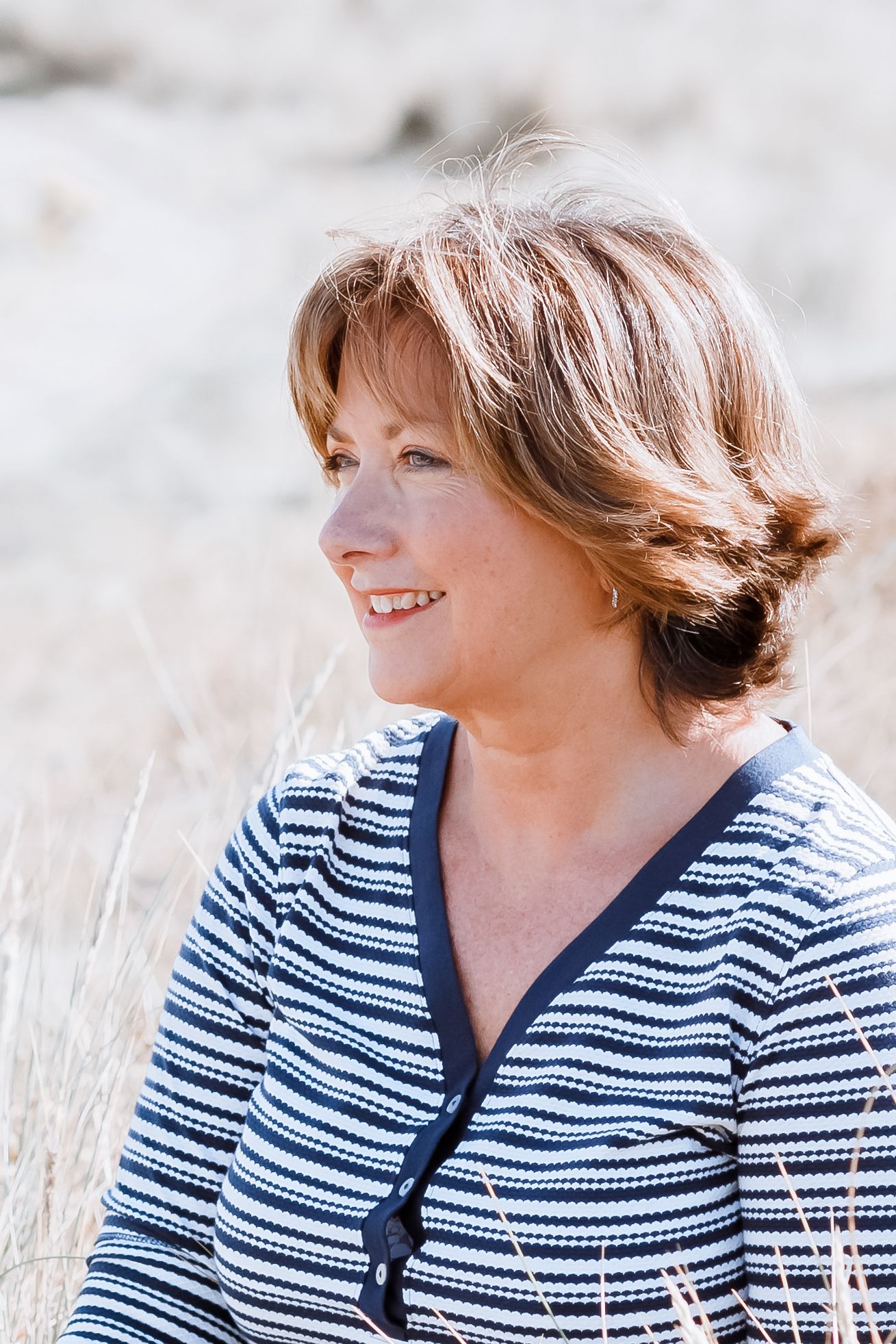 A person in a striped navy and white cardigan sweater smiles while sitting outdoors among tall grass.