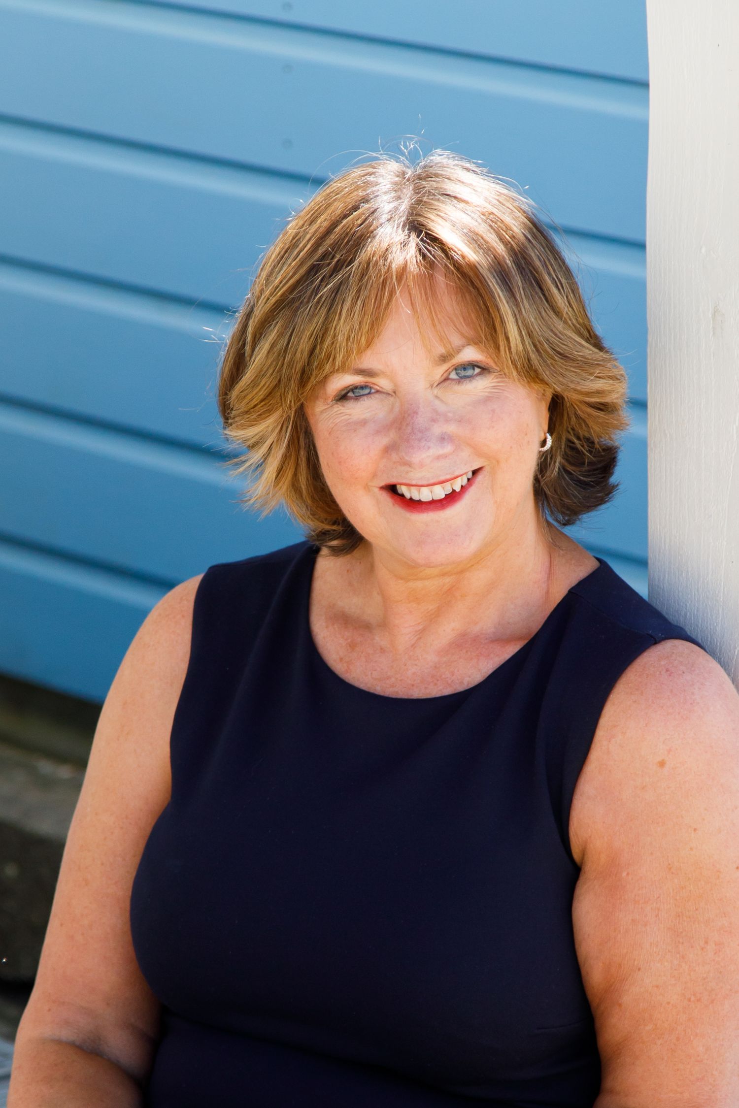 Portrait of someone in a navy sleeveless top smiling warmly against a blue wall background outdoors.