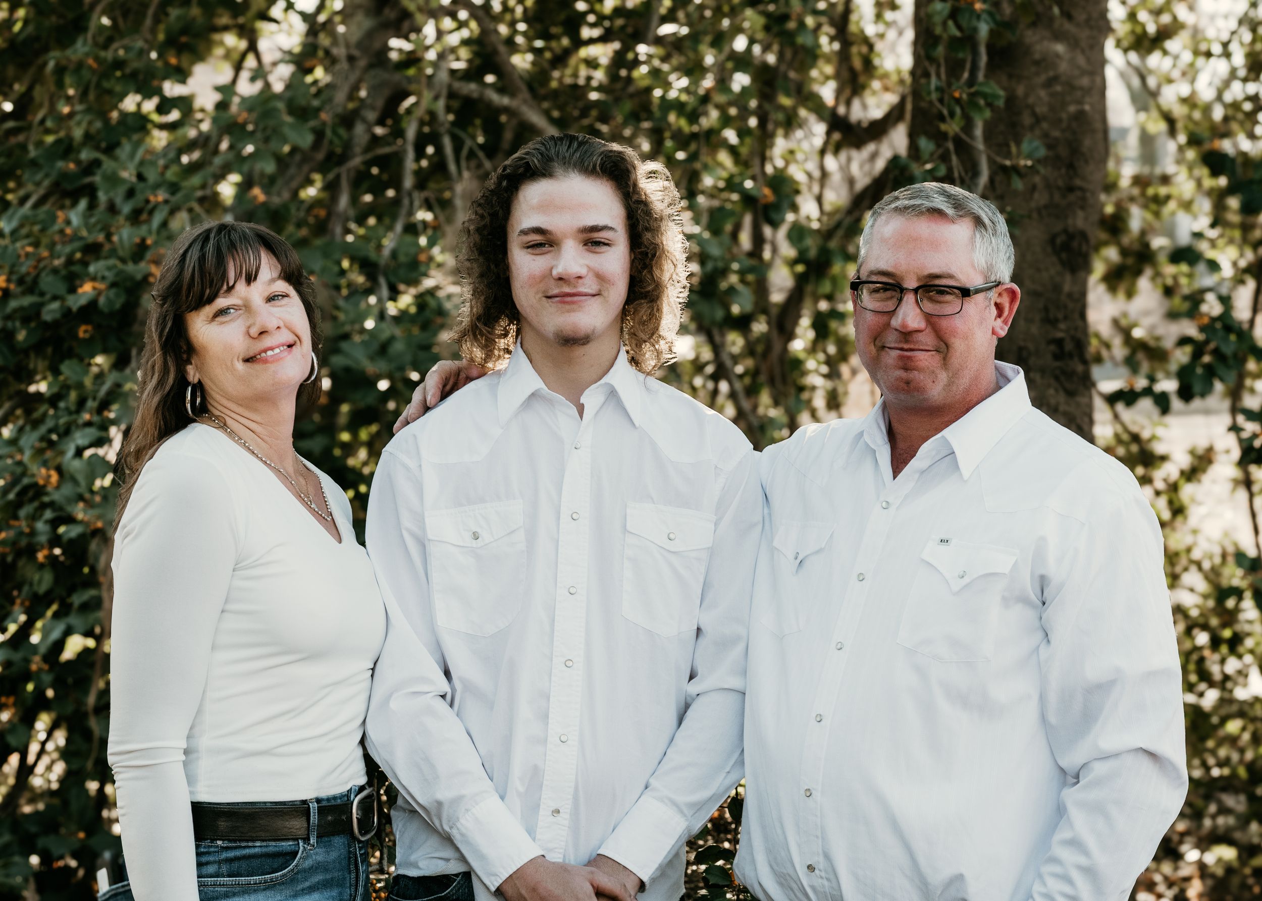A family in matching white shirts poses together outdoors in a natural setting for a casual photo session.