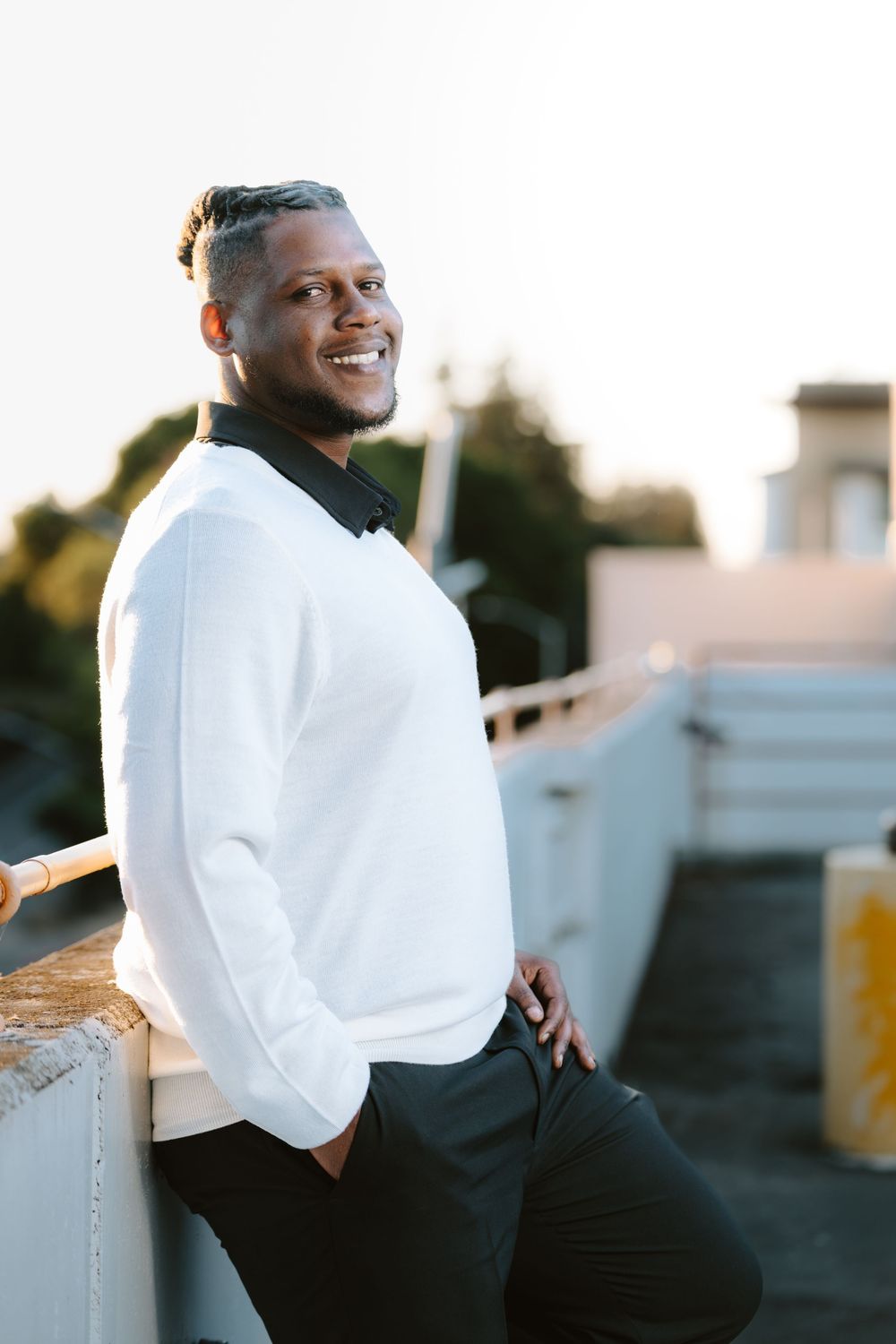 A person in a white sweater and black pants leans against a railing during golden hour outdoors.