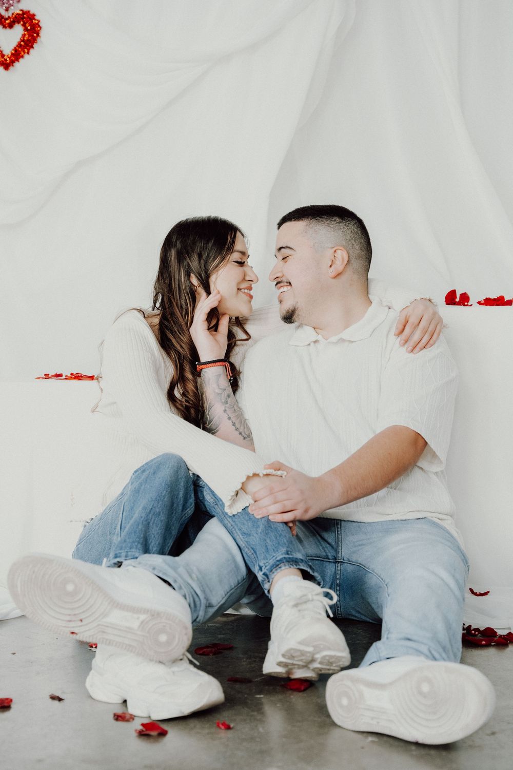 Happy couple in white tops and jeans sitting together on floor with red rose petals in Valentine's Day themed setting.