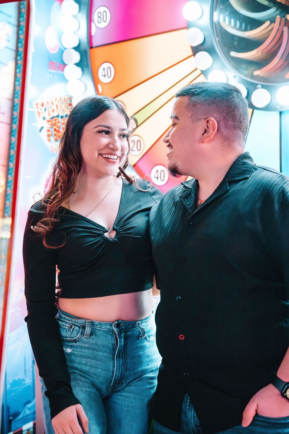 Couple smiling at each other in front of colorful neon lights at night wearing black tops and blue jeans.