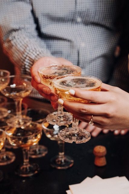Hands holding champagne glasses over a table filled with coupe glasses in warm lighting.