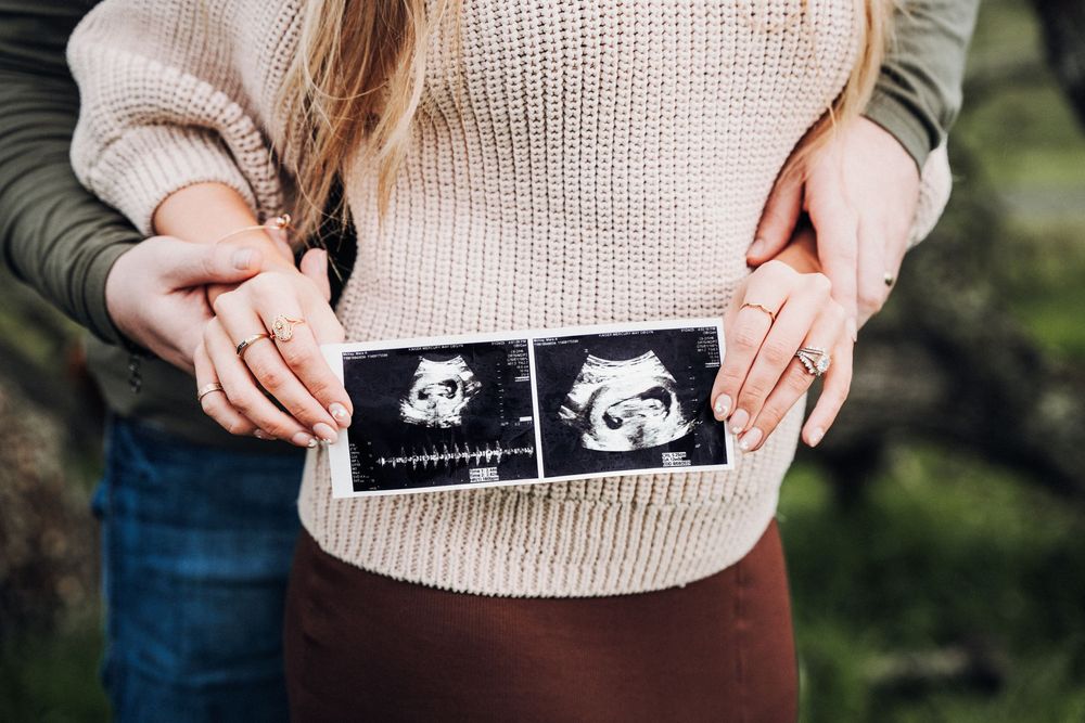 Person wearing beige sweater holds two ultrasound photos against their stomach in pregnancy announcement photo.