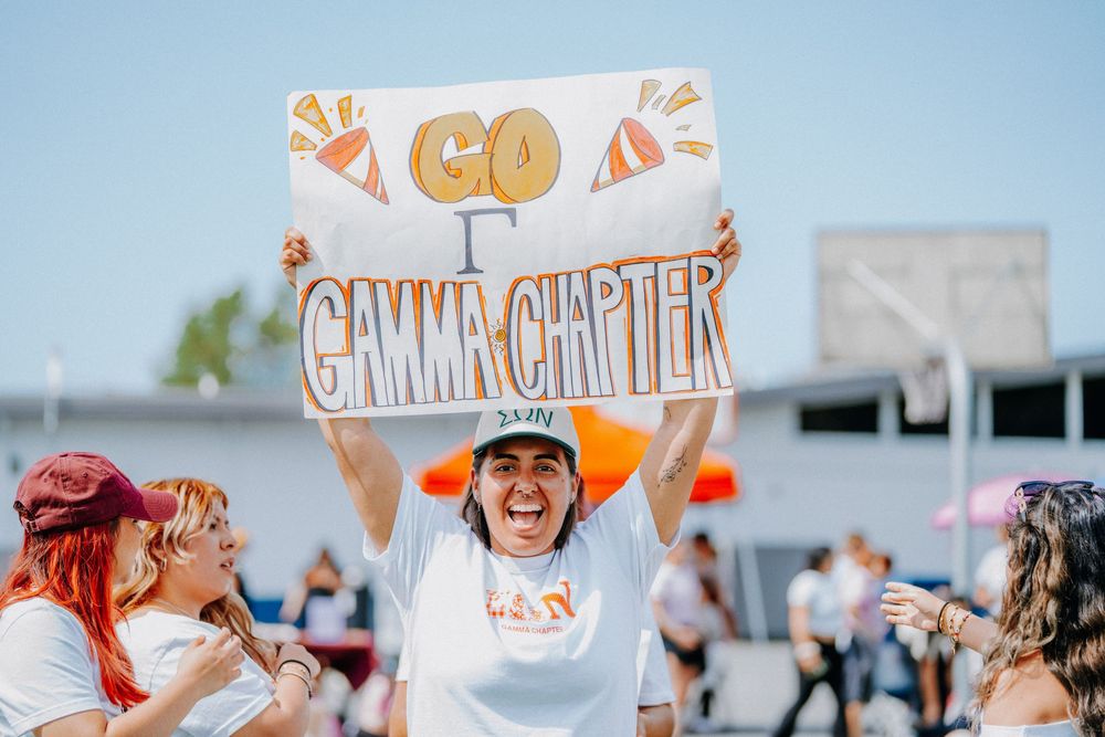 People cheer at an outdoor event with a raised handmade orange and white "Go Gamma Chapter" sign.
