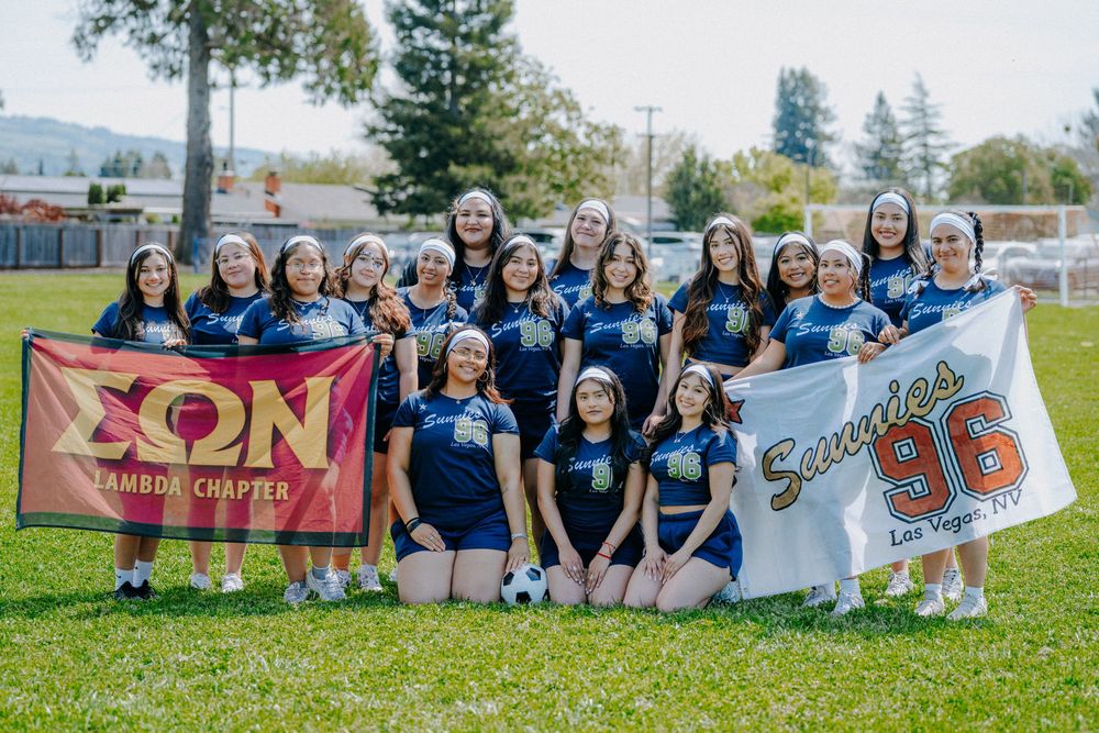 A large group of cheerleaders in navy blue uniforms poses together on grass while holding banners.