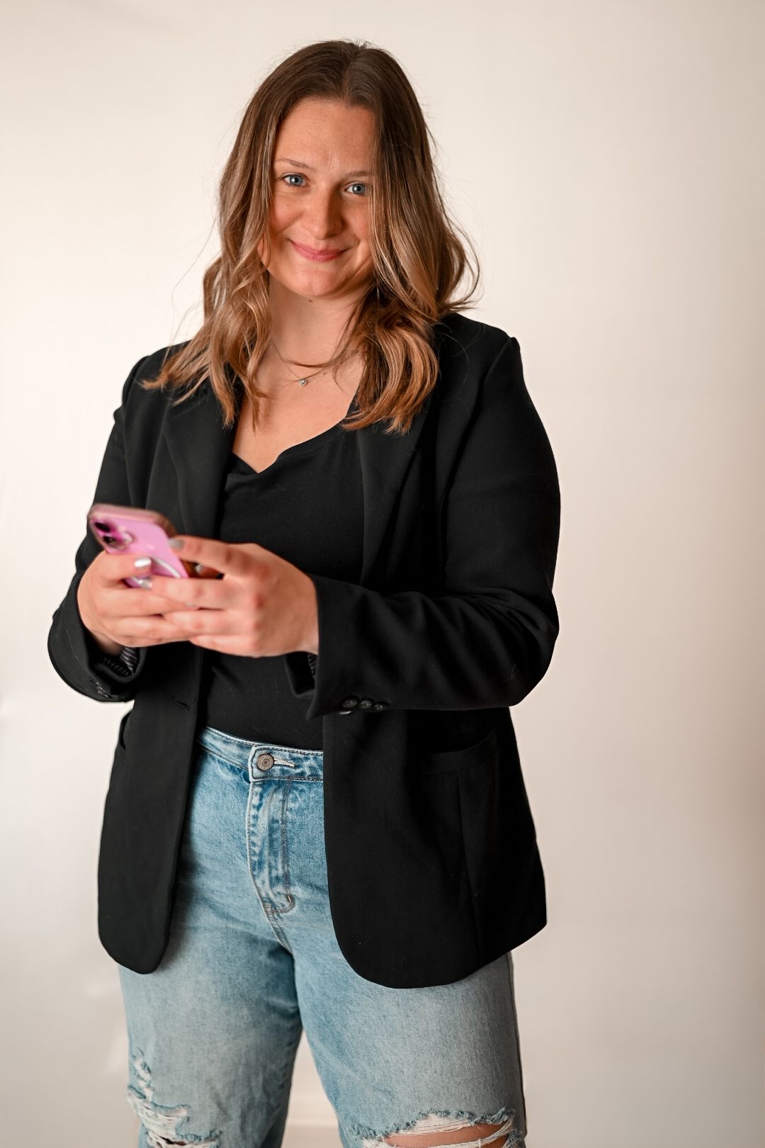 Woman in black blazer and ripped jeans smiling while using a pink smartphone against white background.