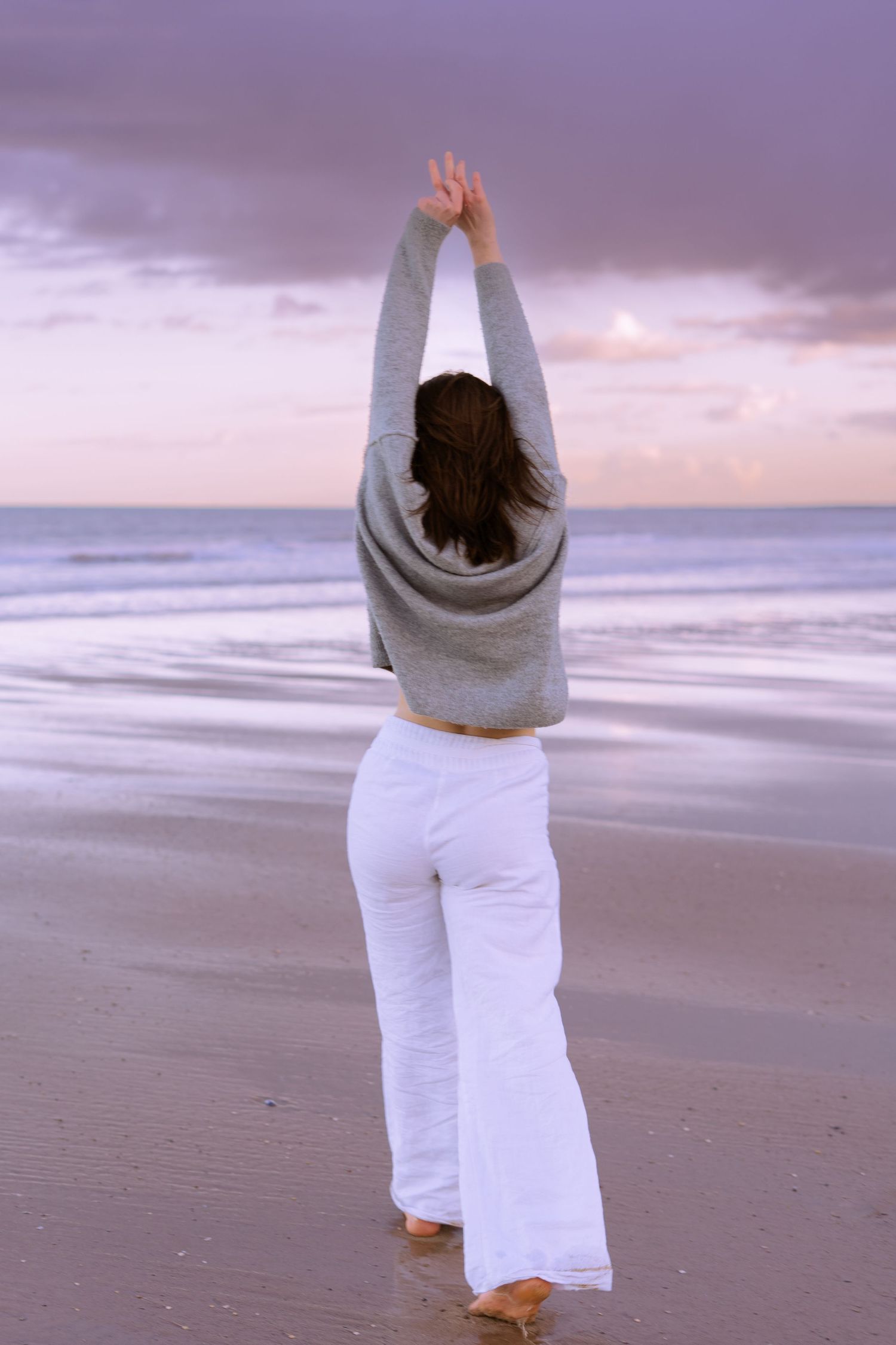 Woman in grey sweater and white pants stretching arms overhead on a serene beach at purple sunset.