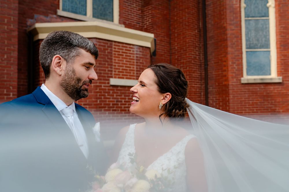 Bride and groom share a romantic glance outside a brick church, her veil flowing beautifully around them.