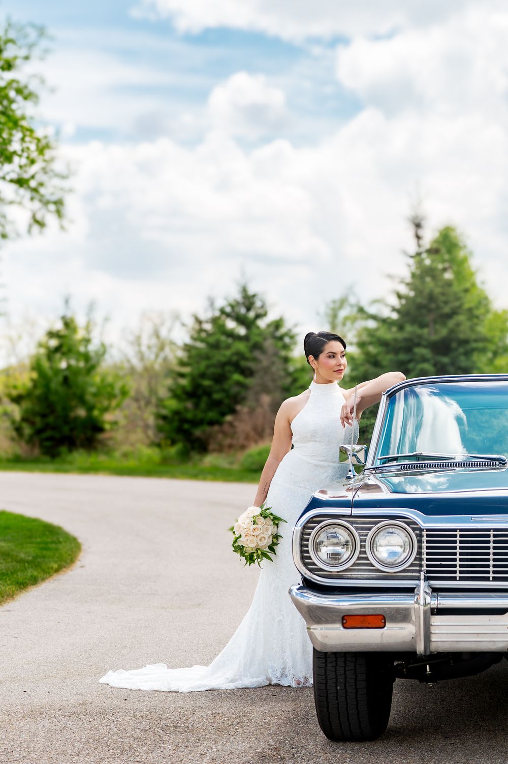 Bride in white lace gown holding bouquet, leaning on classic blue vintage car on a scenic road.