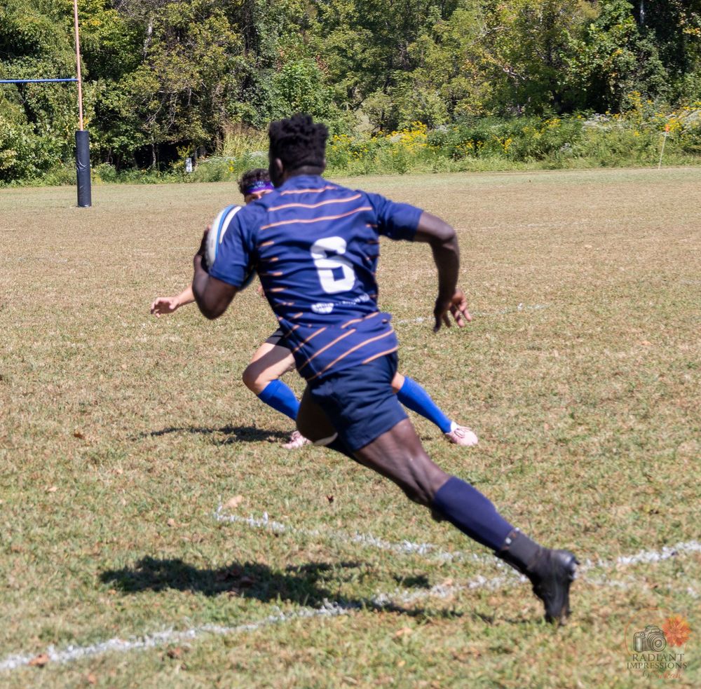 Rugby player wearing number 6 sprints with the ball across a grass field during a match.