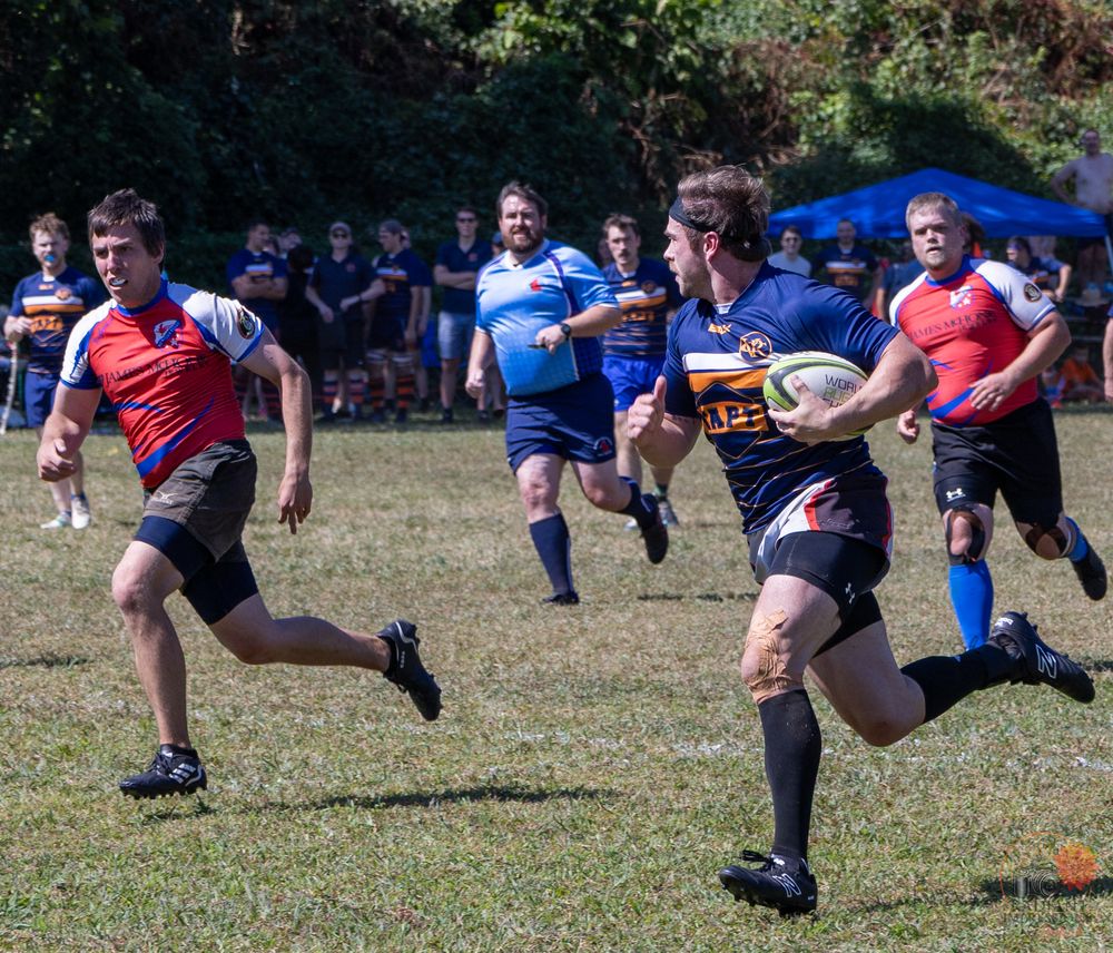 Rugby players compete on a grass field during an outdoor match, with one player carrying the ball while others give chase.