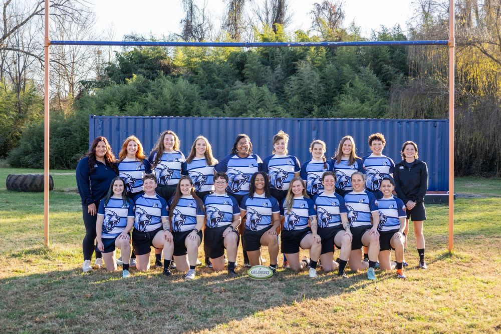 Women's rugby team poses for a group photo in blue and white uniforms on an outdoor field.