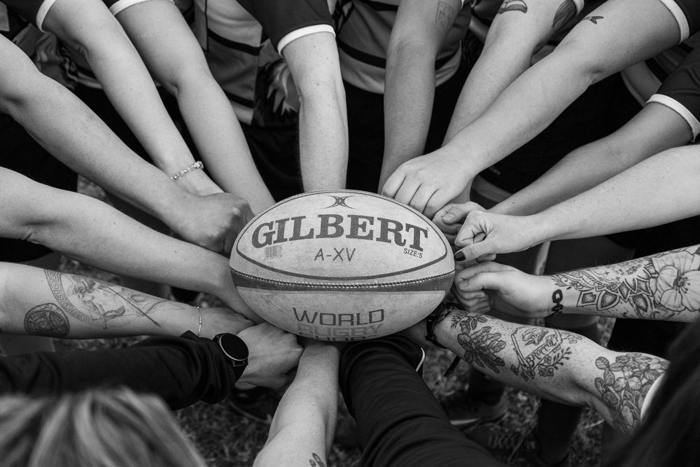 Rugby players form a circle with hands on a Gilbert A-XV rugby ball in a black and white photo.