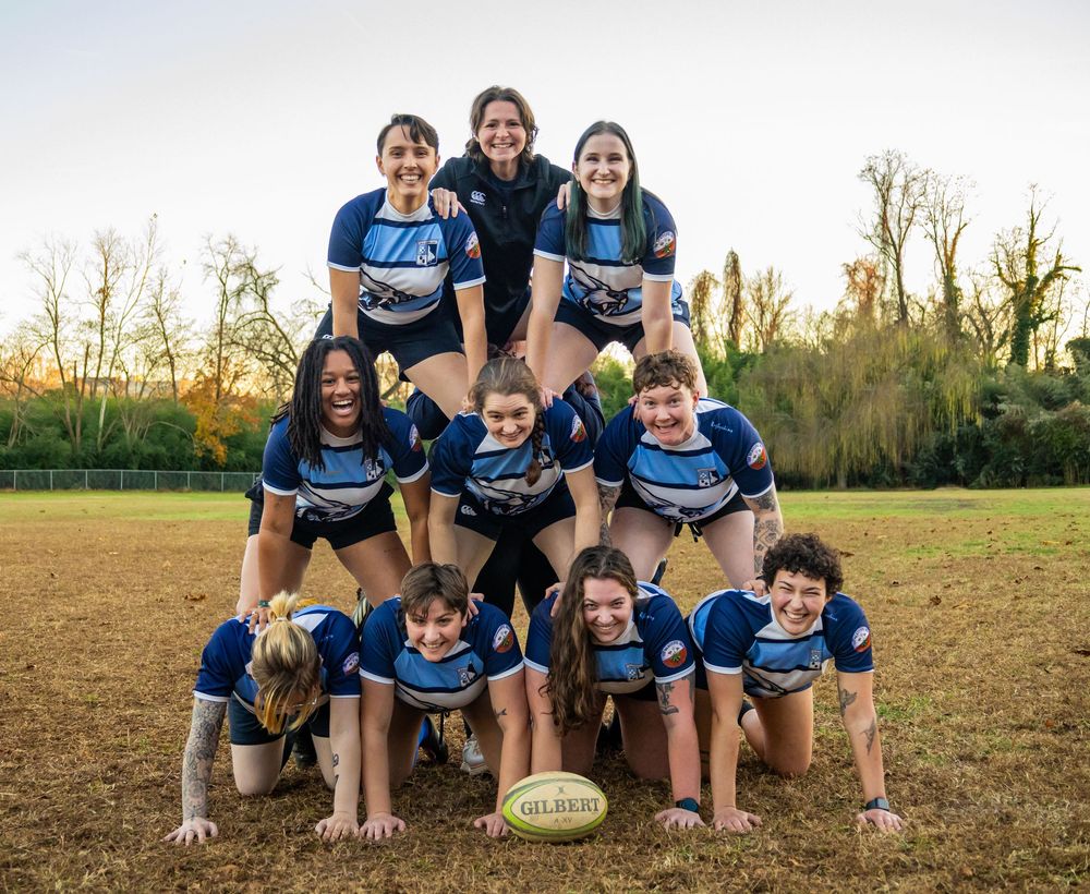 Women's rugby team poses in a fun pyramid formation on a grassy field at sunset.