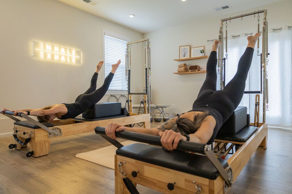 Two women perform Pilates leg exercises on wooden reformer machines in a modern studio with mirrored walls.