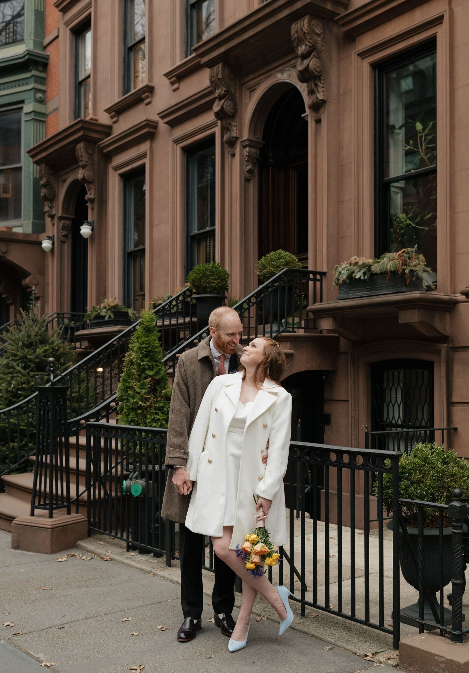 A couple in white outfits walking along a row of classic brownstone buildings in an urban neighborhood.
