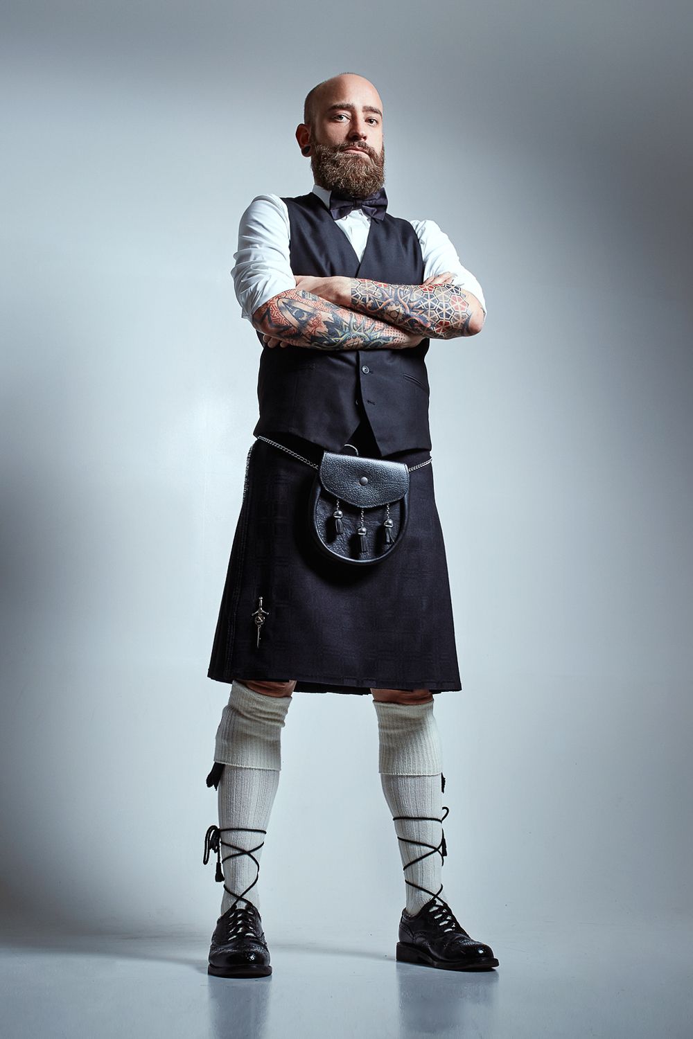 Bald bearded man with tattoos wearing a traditional Scottish kilt outfit with vest and bow tie.
