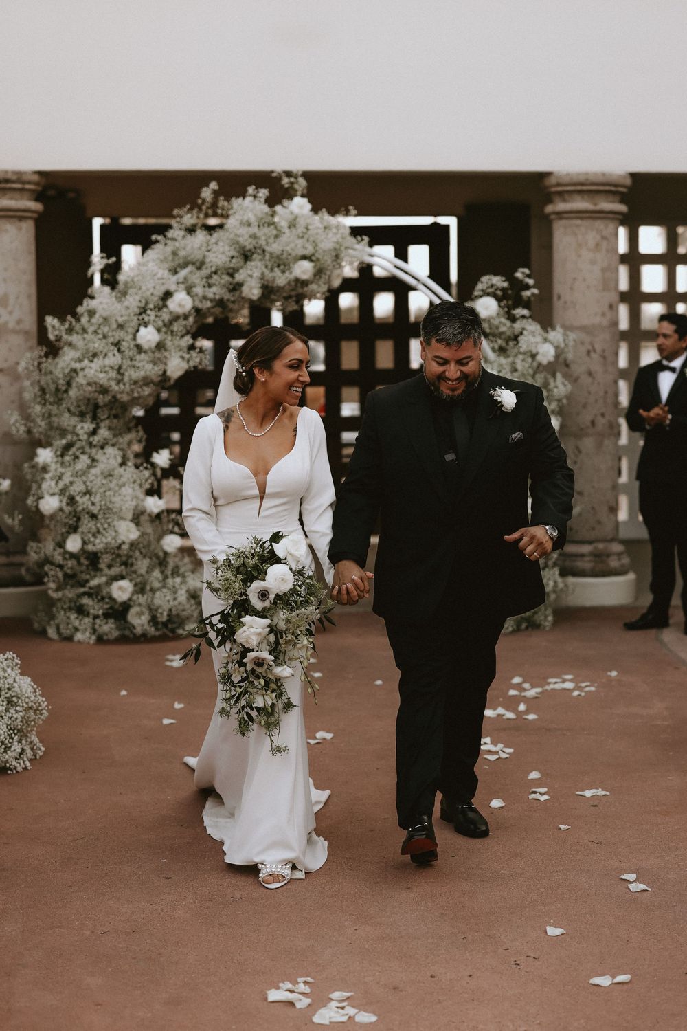 A couple walks through scattered flower petals under an elegant archway decorated with white flowers.