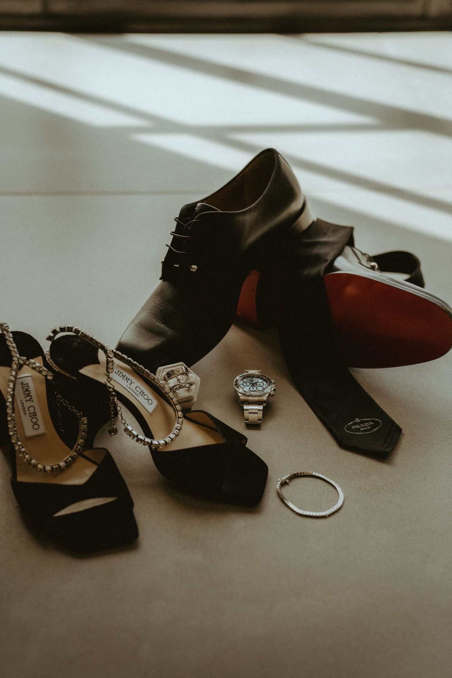 Wedding details including dress shoes, rings, and accessories arranged in dramatic lighting.