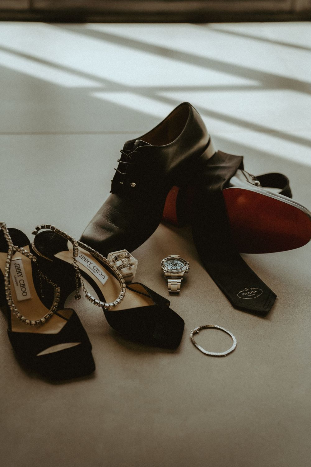 Wedding details including dress shoes, rings, and accessories arranged in dramatic lighting.