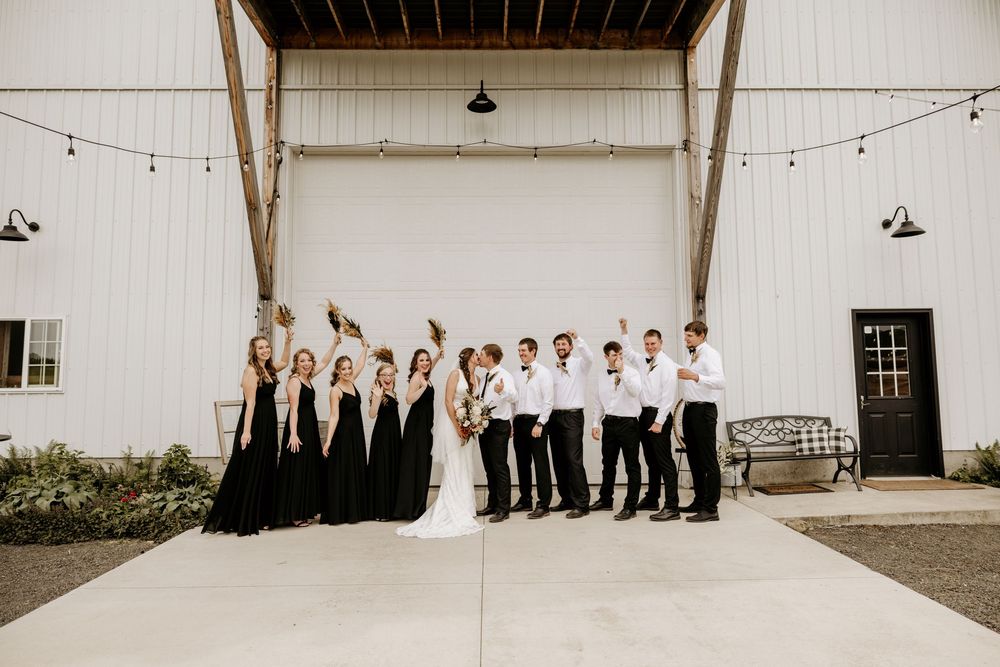 Wedding party lineup against white barn wall with string lights overhead.