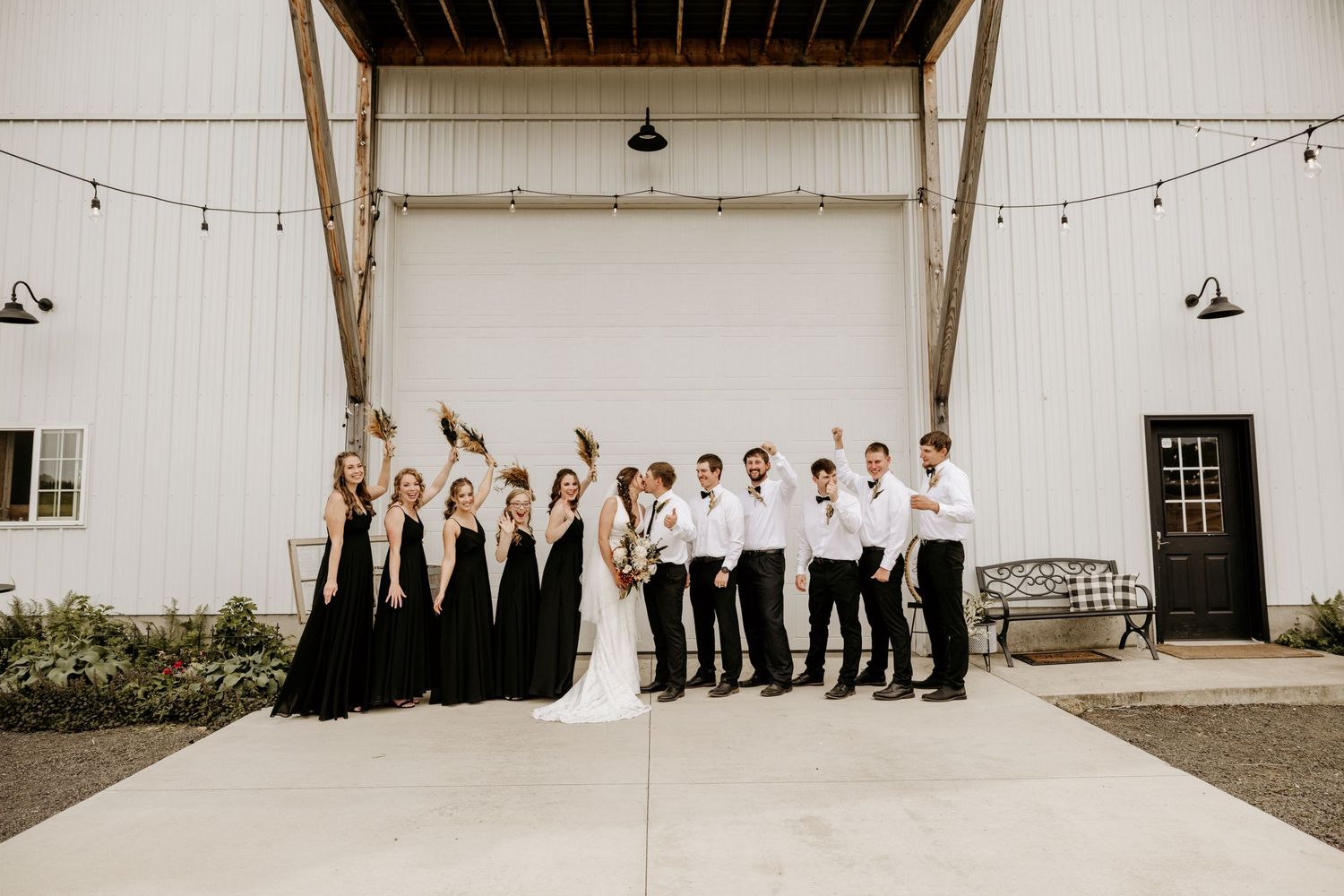 Wedding party lineup against white barn wall with string lights overhead.
