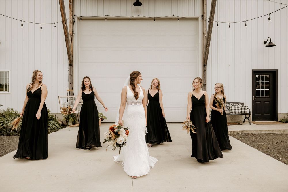 Wedding party poses in front of rustic white barn wall with string lights.
