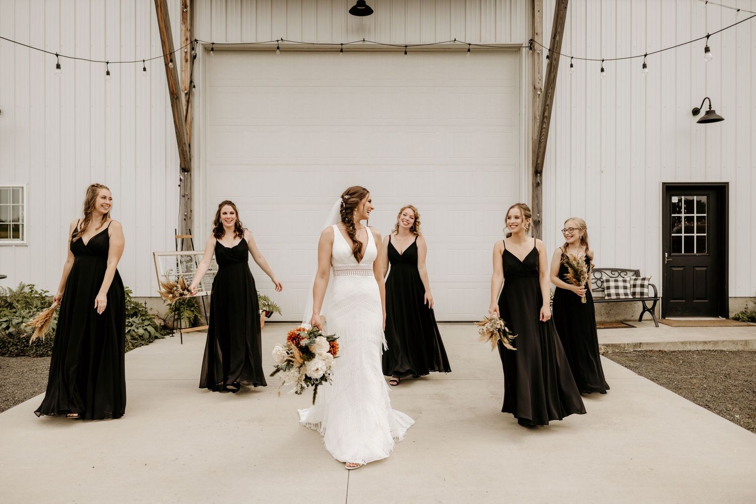Wedding party poses in front of rustic white barn wall with string lights.