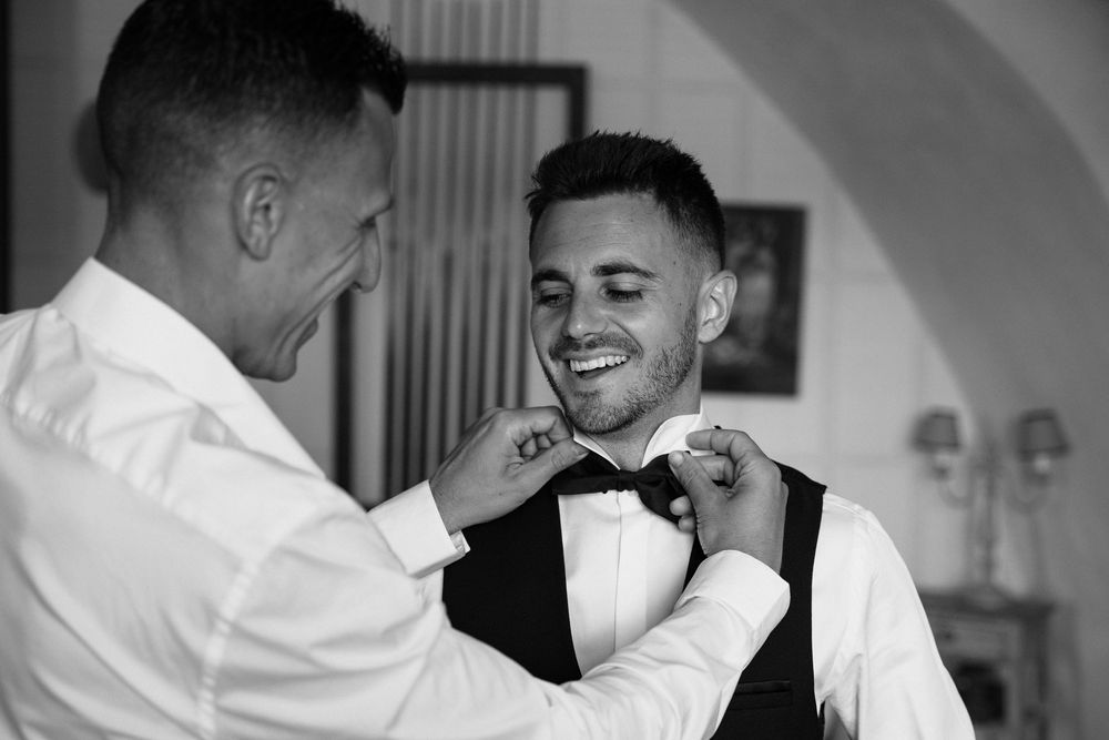 Black and white photo of someone helping adjust a bowtie on a person wearing a vest and formal attire.