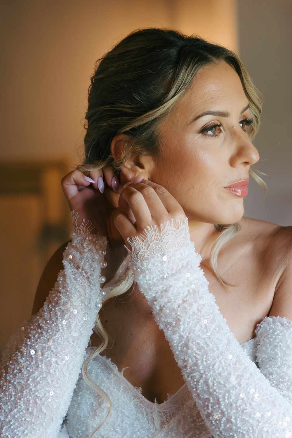 A bride in a long-sleeved lace wedding dress poses in soft window light with a delicate profile view.