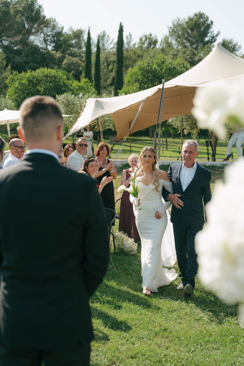 A wedding celebration unfolds under white canopy tents in a lush garden setting with cypress trees in the background.