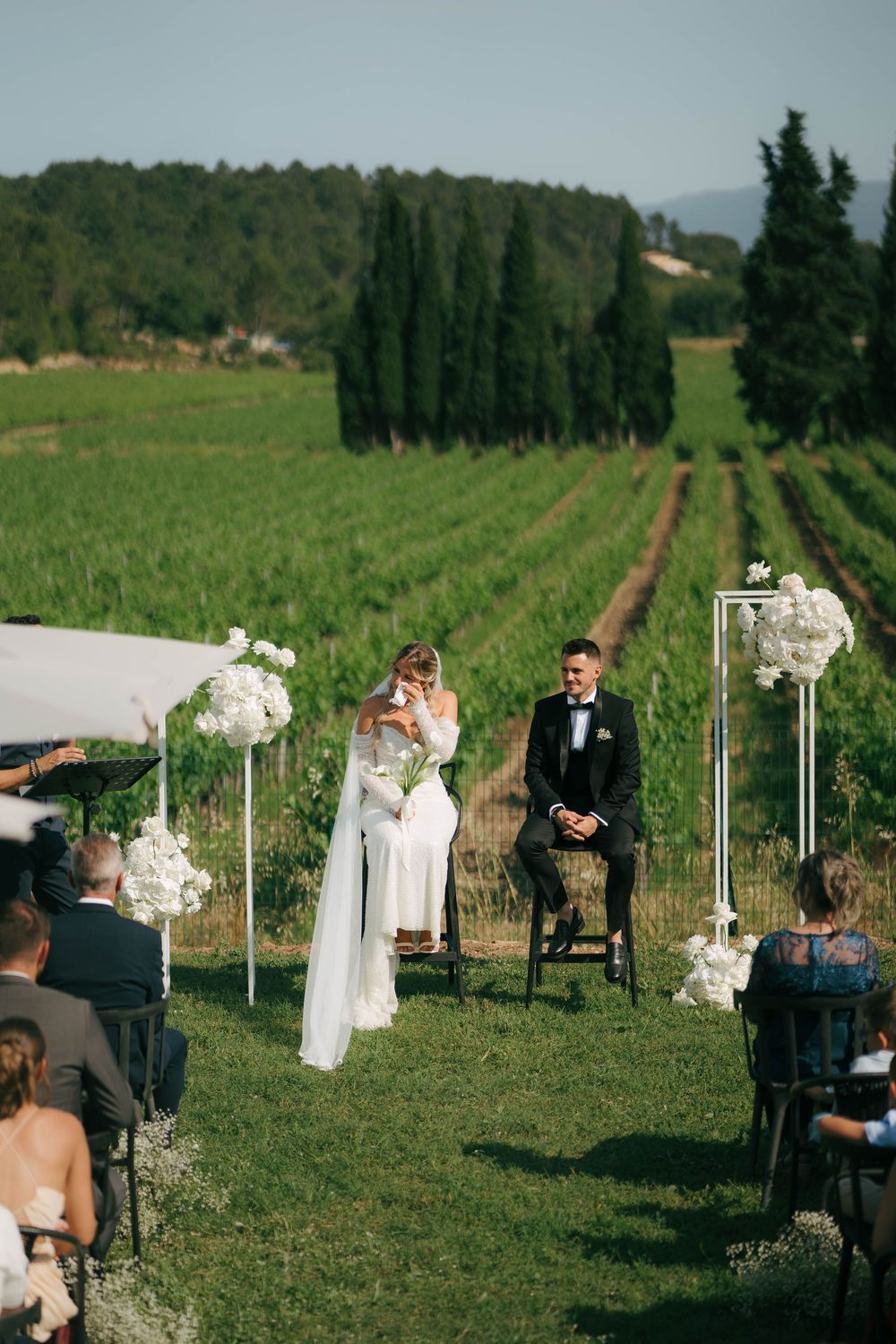 Outdoor wedding ceremony in a vineyard with elegant white floral arrangements and rows of cypress trees in background.