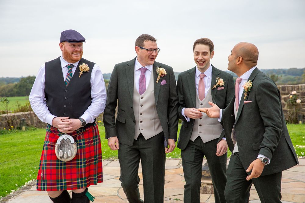 Four groomsmen in formal attire including one in a traditional Scottish kilt chat outdoors before a wedding.