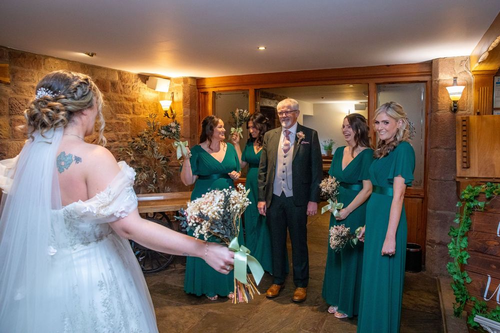 Bride in white gown meets bridesmaids in emerald green dresses and father in a rustic wedding venue.