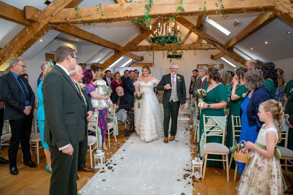 Bride walks down the aisle with her father past guests in a charming barn wedding ceremony venue.