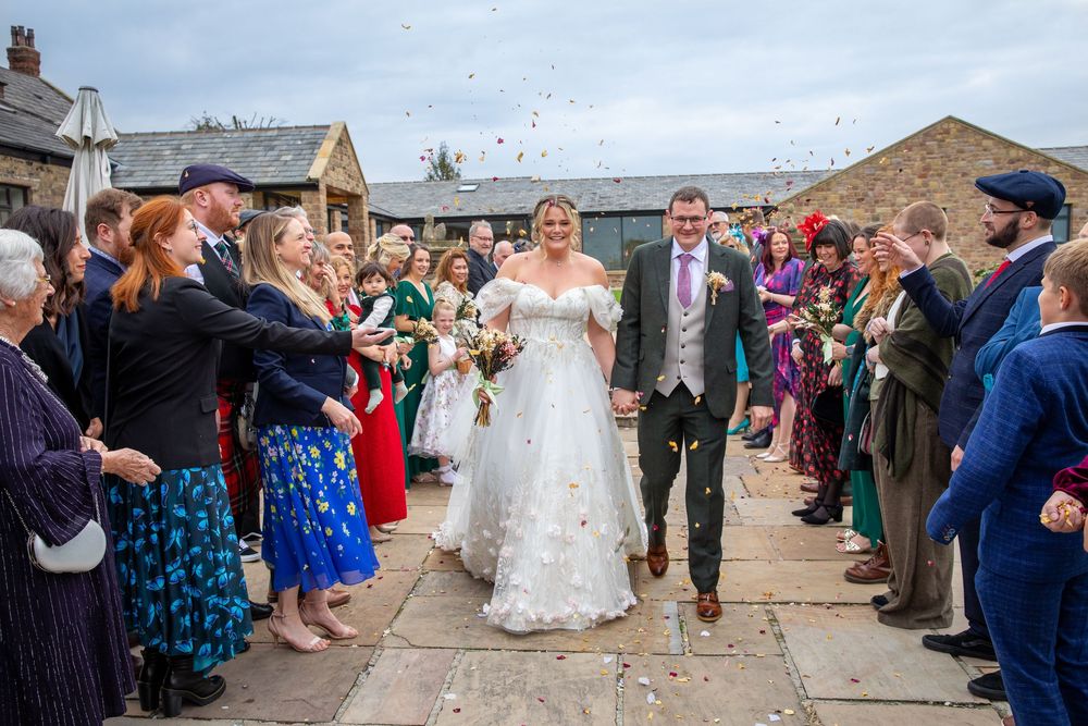 Bride and groom walk through confetti shower surrounded by cheering wedding guests at rustic stone venue.