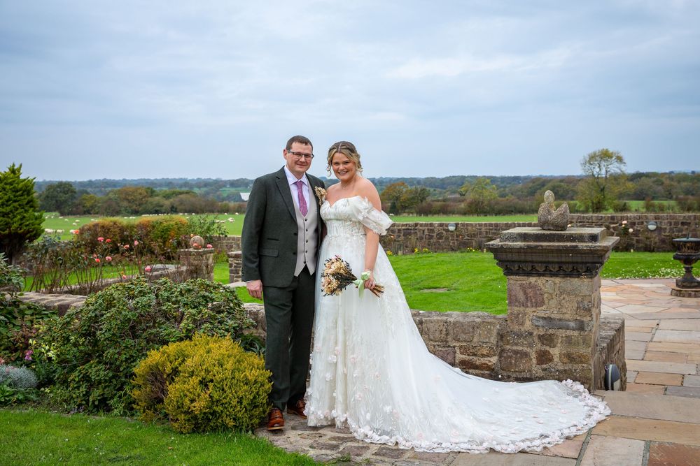 Bride and groom pose together in lace wedding dress and grey suit with scenic countryside views behind.