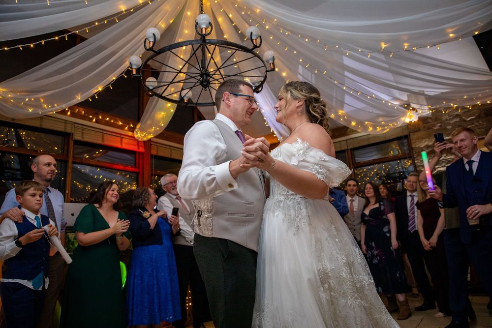 Bride and groom share first dance under fairy lights and chandelier while wedding guests celebrate around them.