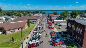 Aerial view of a small-town street festival with classic cars lining the road near a scenic lakefront.