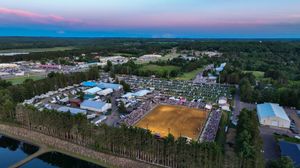 Aerial twilight view of a large outdoor fair with crowds, colorful tents, sandy arena, and lush forest surroundings.