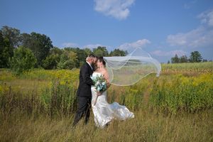 Bride and groom share a romantic kiss in a sunny meadow, her veil flowing in the breeze, surrounded by wildflowers.