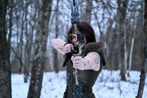 Woman in medieval costume drawing a bow and arrow in a snowy winter forest setting.