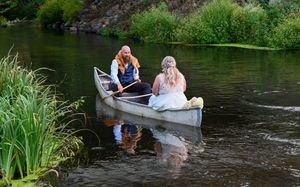 Bride and groom in a canoe paddling along a scenic river surrounded by green vegetation.