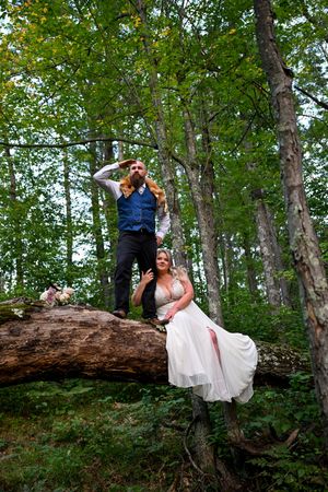 Bride in white gown and groom in blue vest pose on a fallen log in a lush green forest wedding photo.