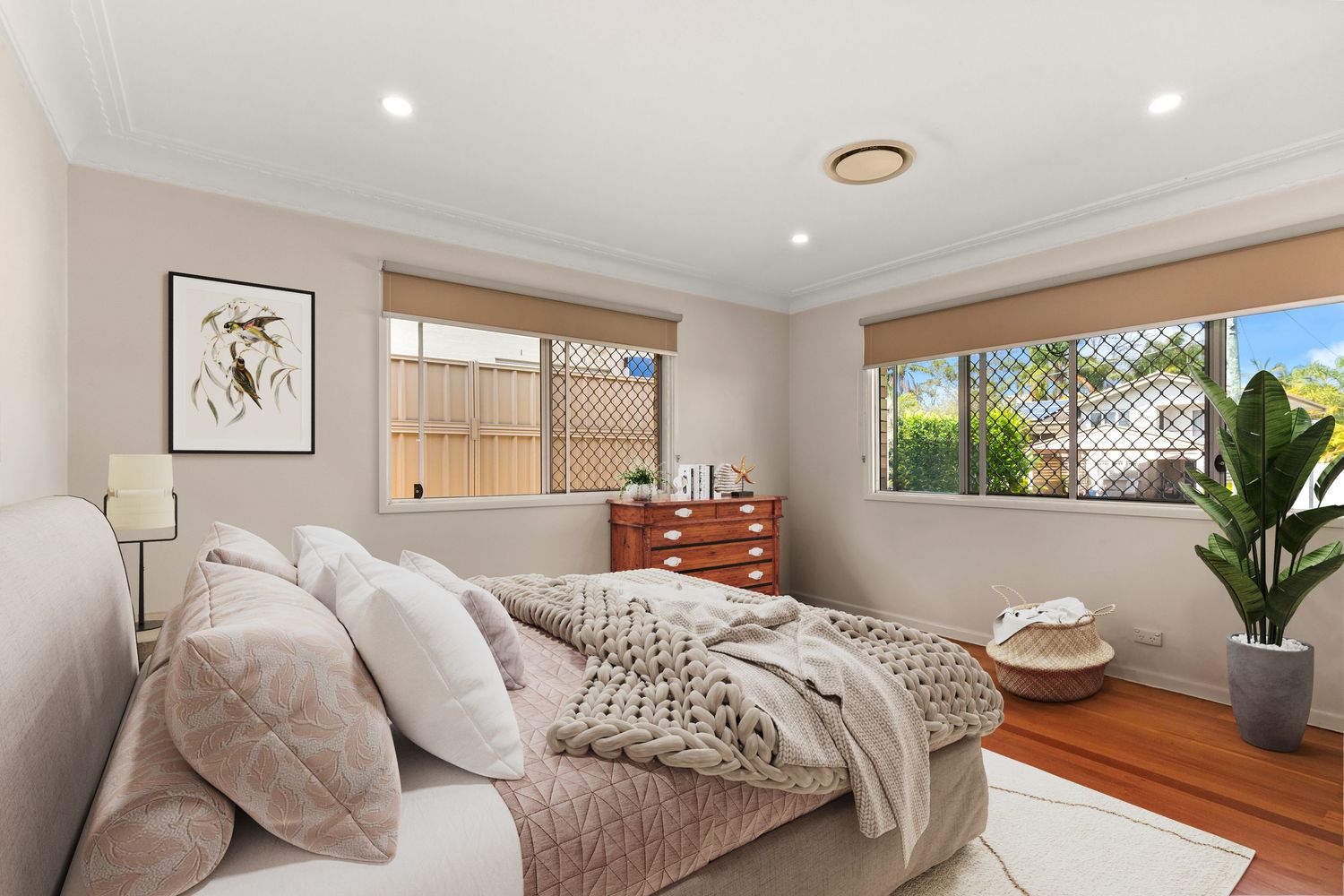 Neutral bedroom with hardwood floors, white bedding, decorative cushions, roller blinds, and indoor plant.