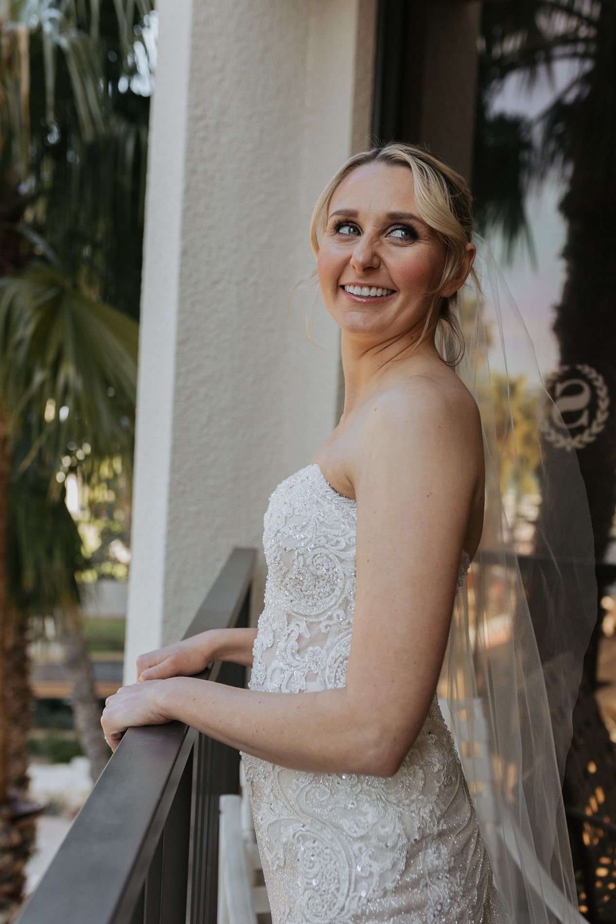 A bride in a white lace wedding dress poses on a balcony with palm trees in the background.