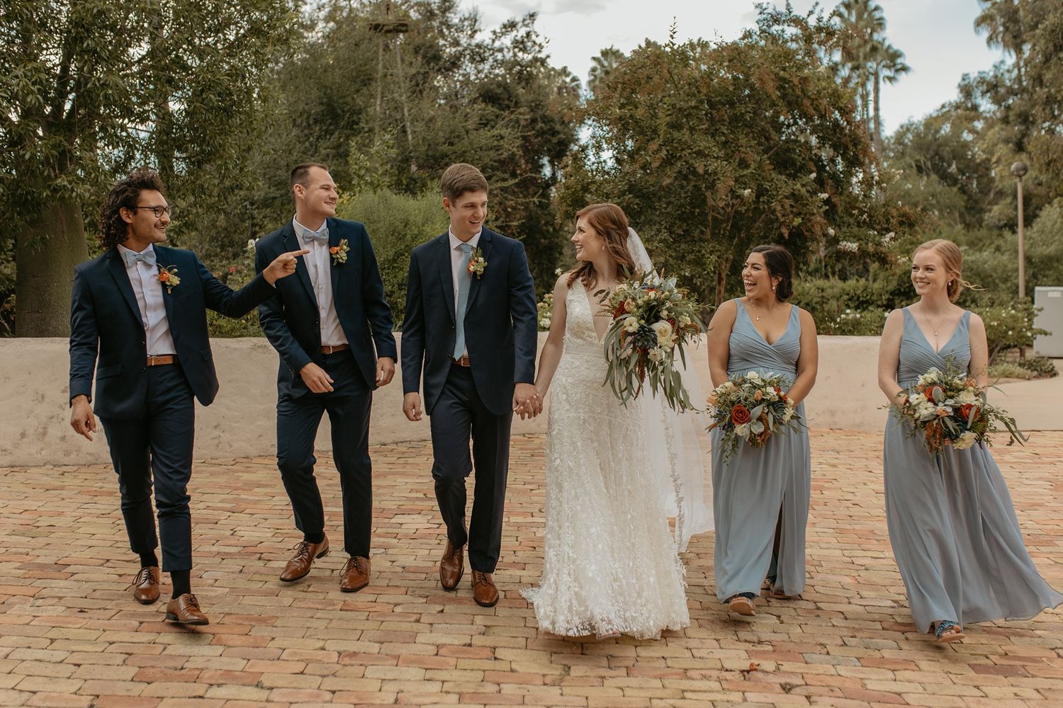 Wedding party walking outdoors, bride in lace gown with bouquet, groom and groomsmen in navy suits, bridesmaids in blue dresses.