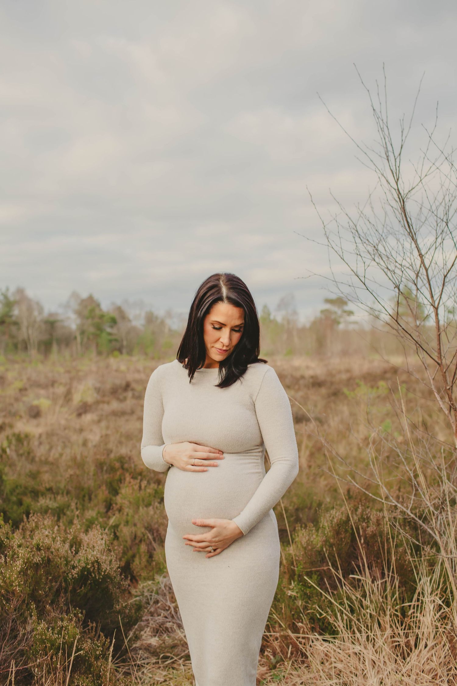 Pregnant woman in gray dress cradling her baby bump outdoors in a natural field setting during maternity photoshoot.